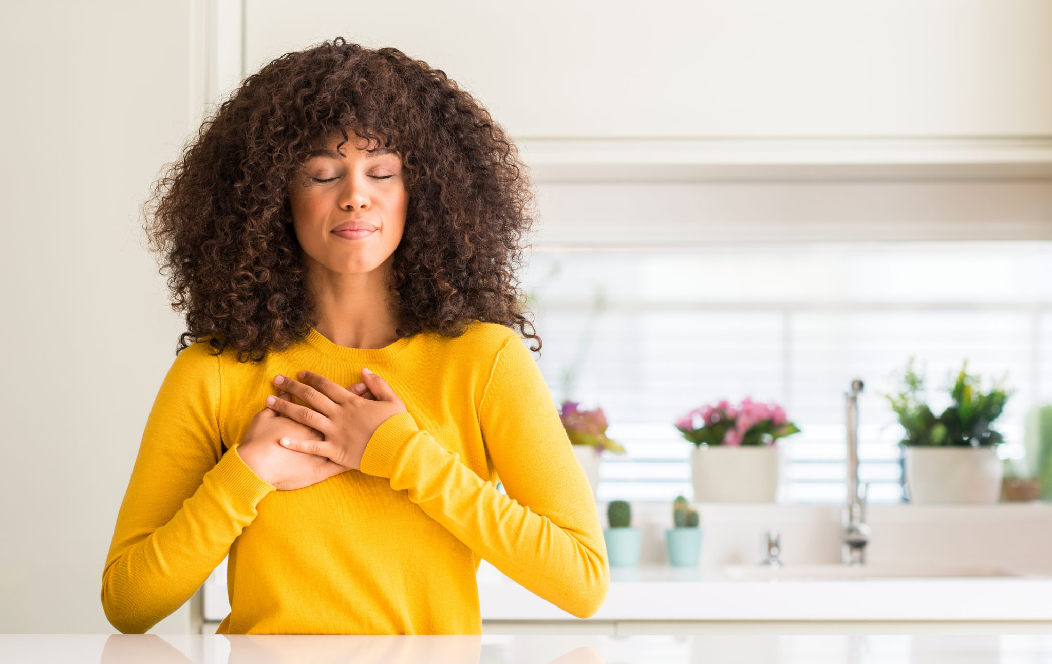 African american woman wearing yellow sweater at kitchen smiling with hands on chest with closed eyes and grateful gesture on face.